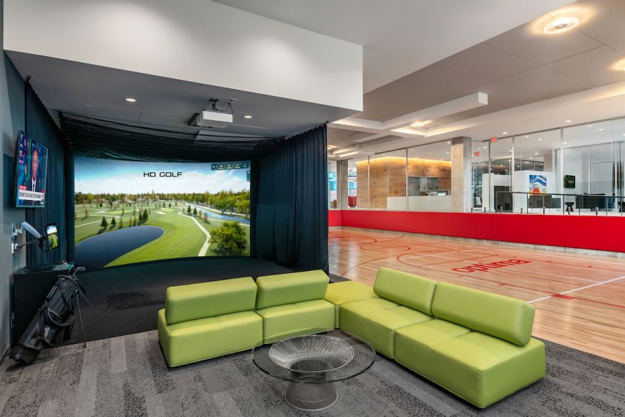 Optima Verdana luxury apartment with Green couches and a glass table near a golf simulator, with a basketball court visible in the background.