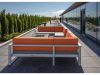 Optima Verdana luxury apartment with Outdoor rooftop patio with an orange sectional sofa, glass fire pit, lounge chairs, and greenery along the edge, under a clear sky.