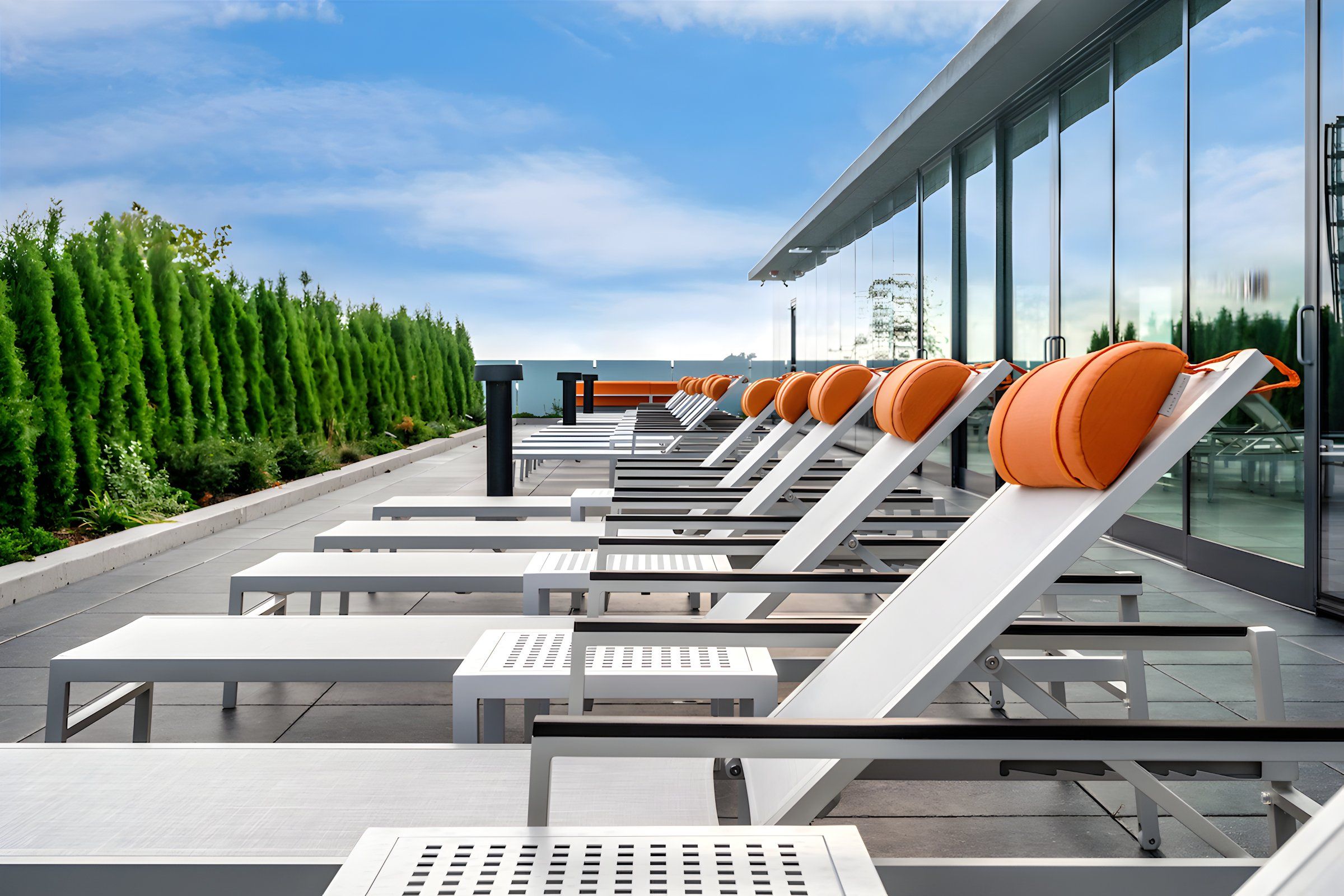 Optima Verdana luxury apartment with Row of white lounge chairs with orange pillows on a rooftop patio beside green shrubs and glass windows.