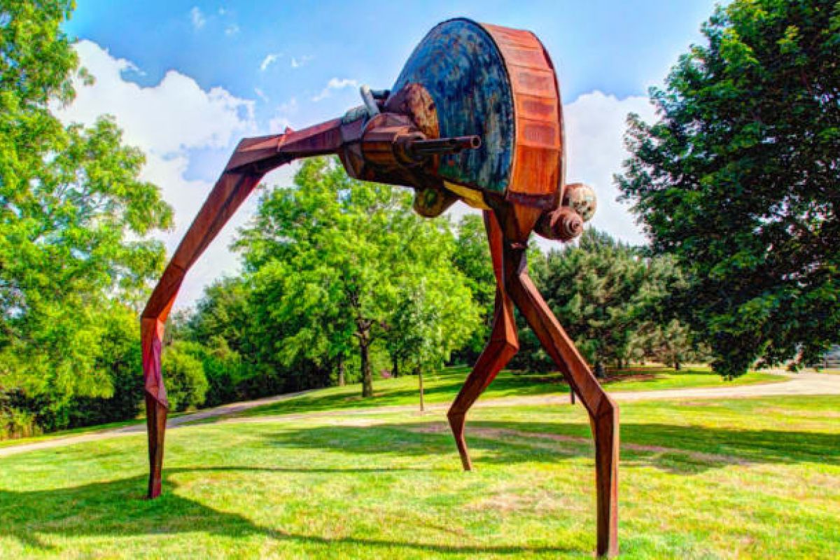 Optima Verdana luxury apartment with Large rusted metal sculpture with three long legs stands on green grass at Skokie’s Northshore Sculpture Park, surrounded by trees and blue sky.