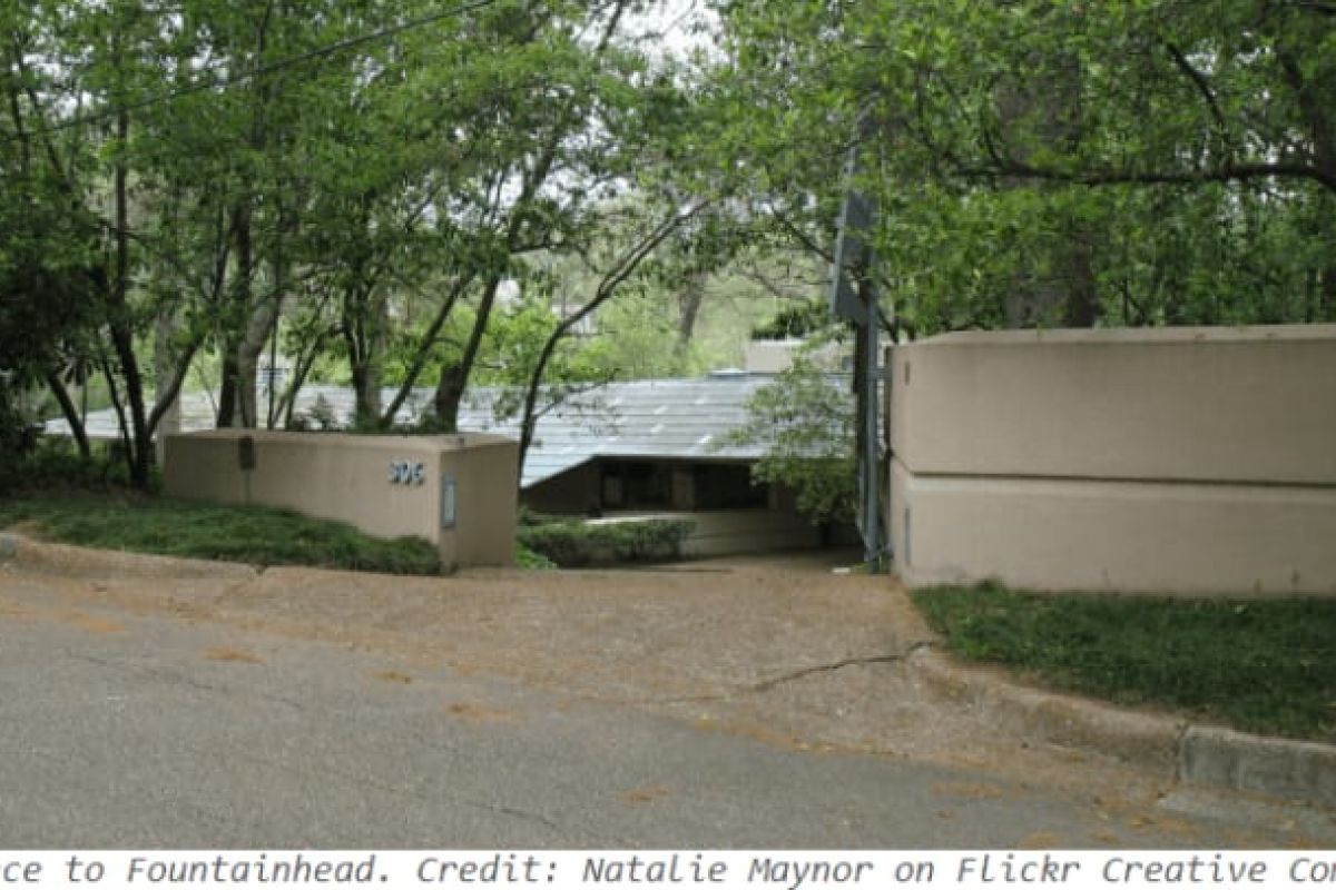 Optima Verdana luxury apartment with Gated entrance to a Forever Modern house surrounded by trees, with beige walls and a sloped driveway leading down.
