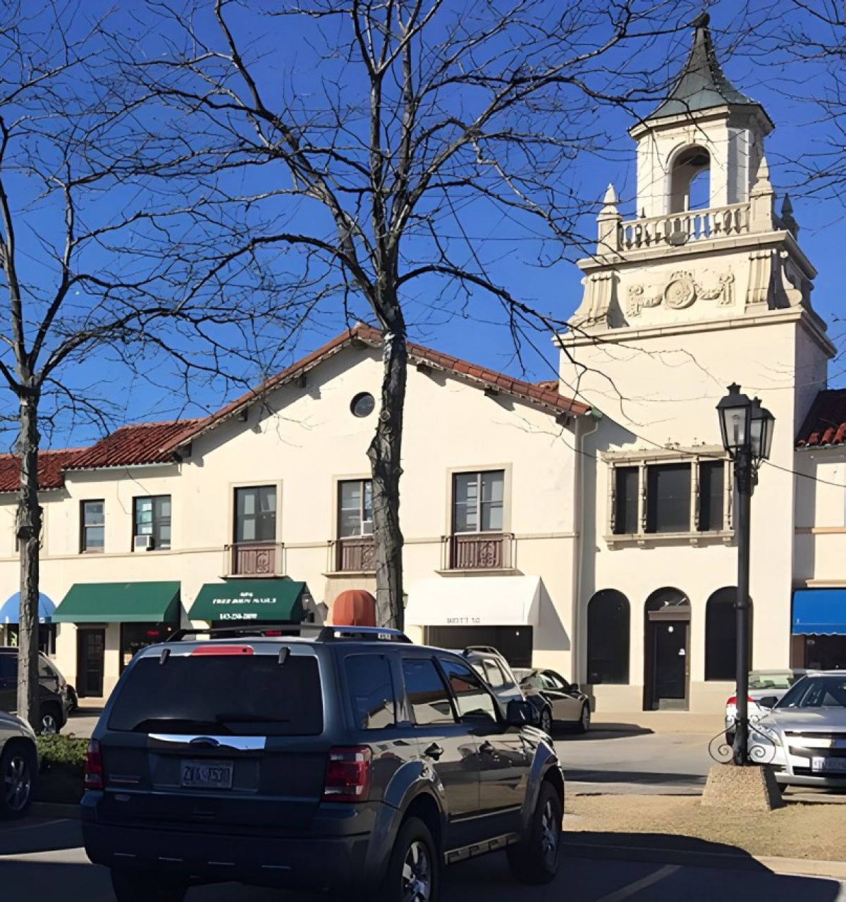 Optima Verdana luxury apartment with Cars parked in front of Spanish-style shops and buildings with a clocktower, under a clear blue sky.