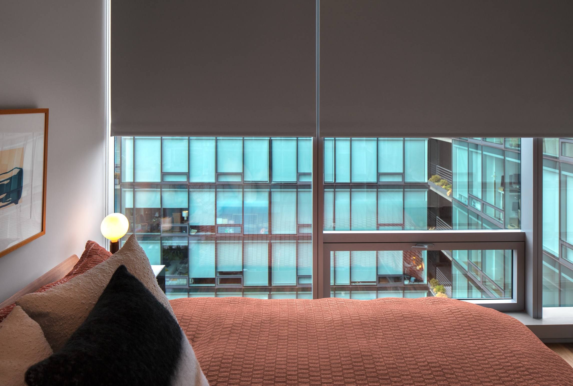 Optima Verdana luxury apartment with Modern bedroom with a made bed, lamp, and large window showing a building with blue-tinted glass panels.