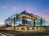 Optima Verdana luxury apartment with Modern glass building from the Optima Portfolio on a city corner, with bright lights, trees, and lively common features at dusk.