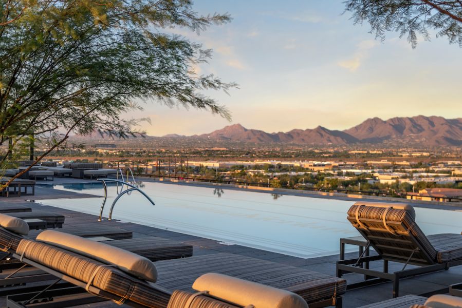 Rooftop infinity pool at Optima Kierland Apartments with lounge chairs, overlooking a cityscape and distant mountains at sunset.
