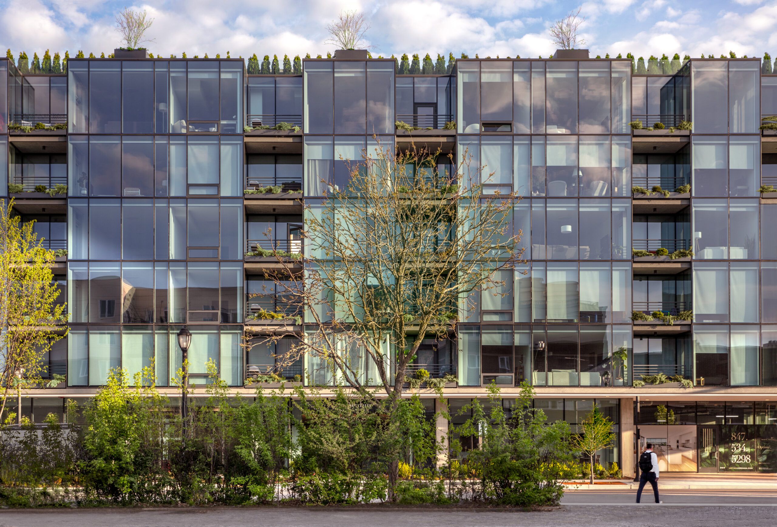 Optima Verdana luxury apartment with Modern glass apartment building, Optima Verdana, with balconies and greenery on the North Shore; a person walks by on a sunny day.
