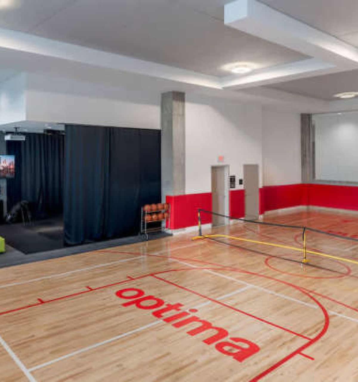 Optima Verdana luxury apartment with Spacious home indoor court with full-court basketball, pickleball net, red and wood floors, green sofa, and a TV in the corner.