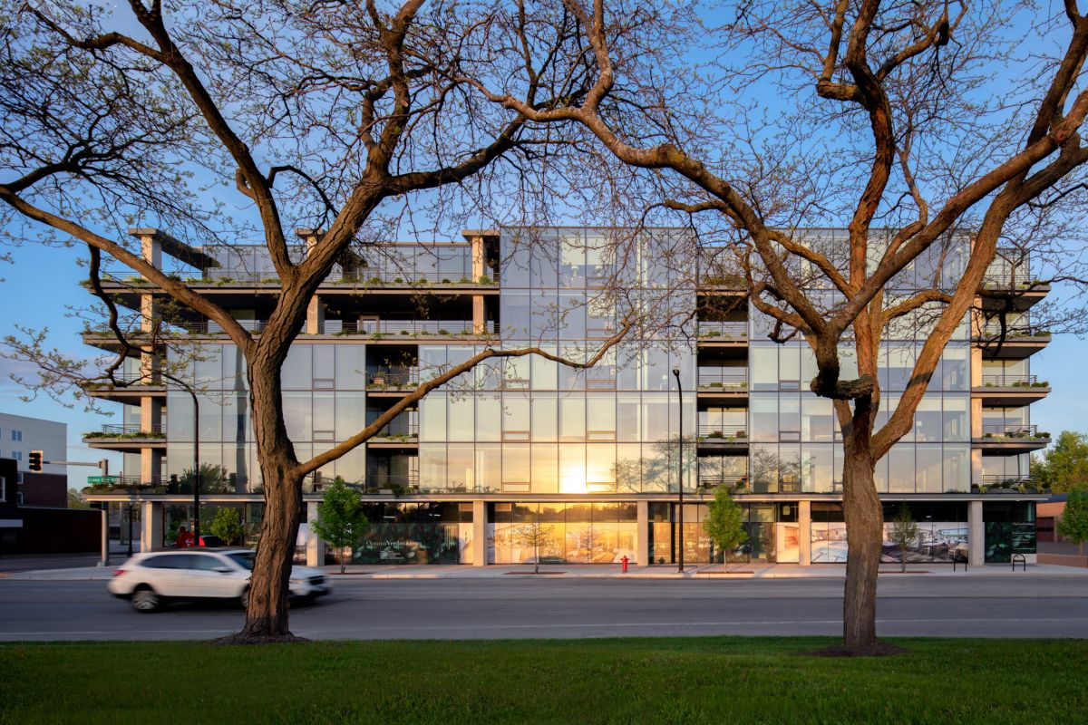 Optima Verdana luxury apartment with Modern glass apartment building reflected in evening light, framed by two leafless trees and a car driving by.