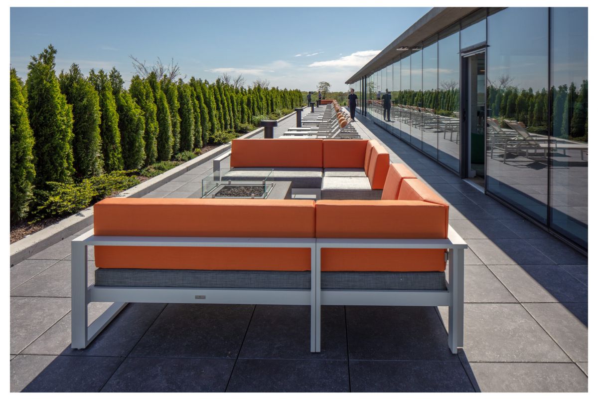 Optima Verdana luxury apartment with Outdoor rooftop patio with an orange sectional sofa, glass fire pit, lounge chairs, and greenery along the edge, under a clear sky.