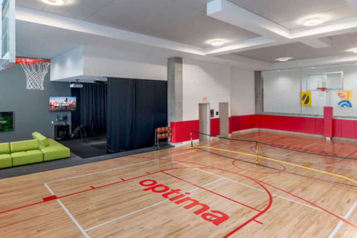 Optima Verdana luxury apartment with Spacious home indoor court with full-court basketball, pickleball net, red and wood floors, green sofa, and a TV in the corner.