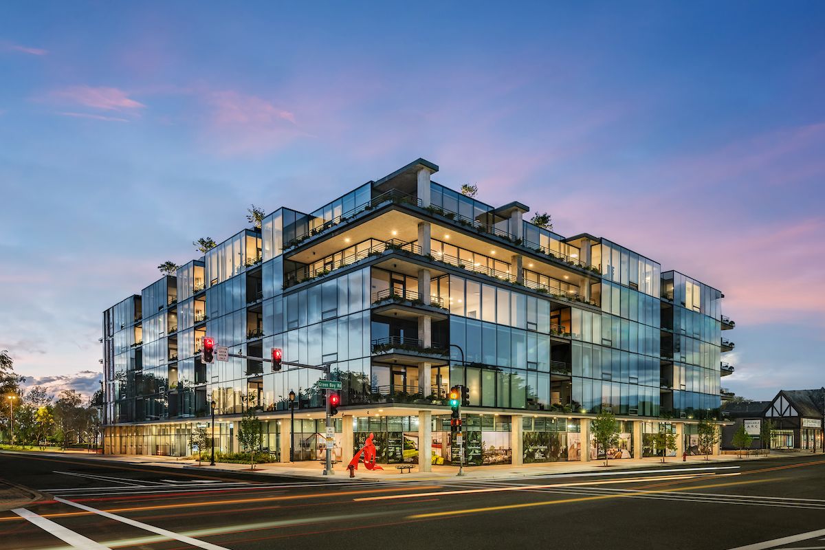 Optima Verdana luxury apartment with Modern glass building from the Optima Portfolio on a city corner, with bright lights, trees, and lively common features at dusk.