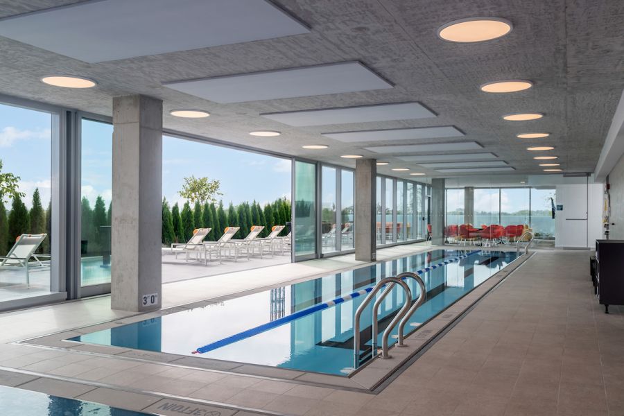 Optima Verdana luxury apartment with Indoor pool with metal handrails, large windows, and outdoor lounge chairs visible through the glass.
