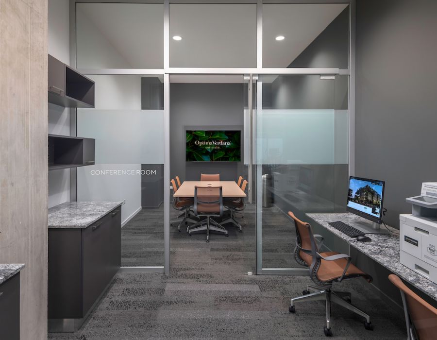 Optima Verdana luxury apartment with Modern office with a glass-walled conference room, chairs, computers, and a printer on a gray carpeted floor.