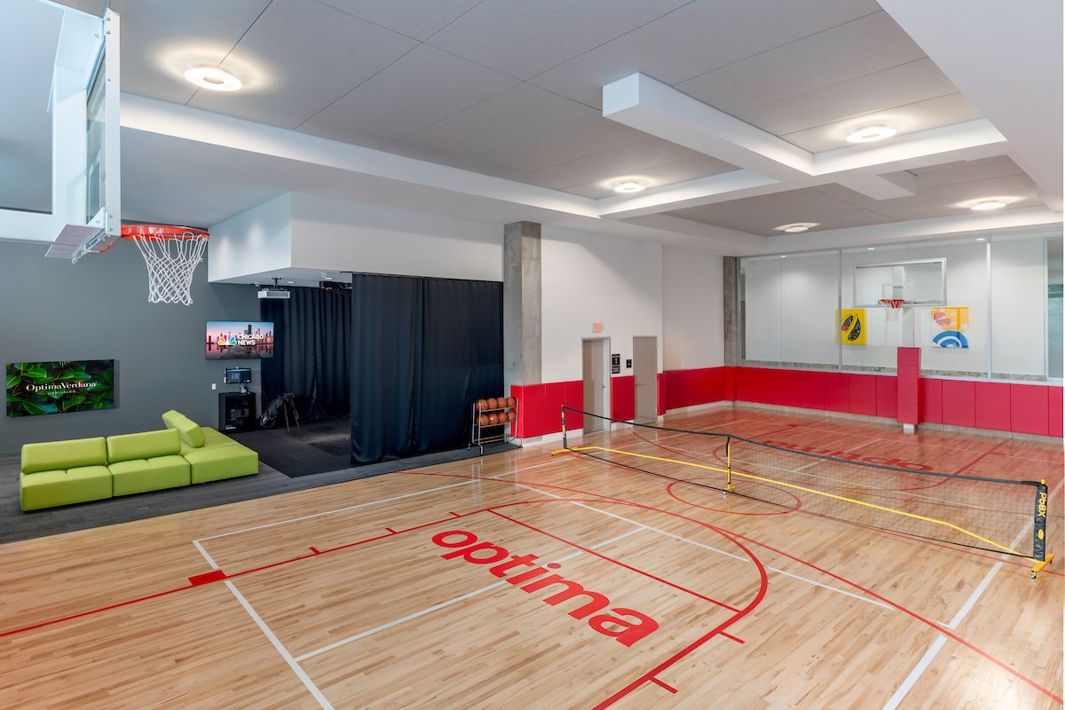 Optima Verdana luxury apartment with Indoor gym with basketball court, small tennis net, green couch, TV, and red and white walls.