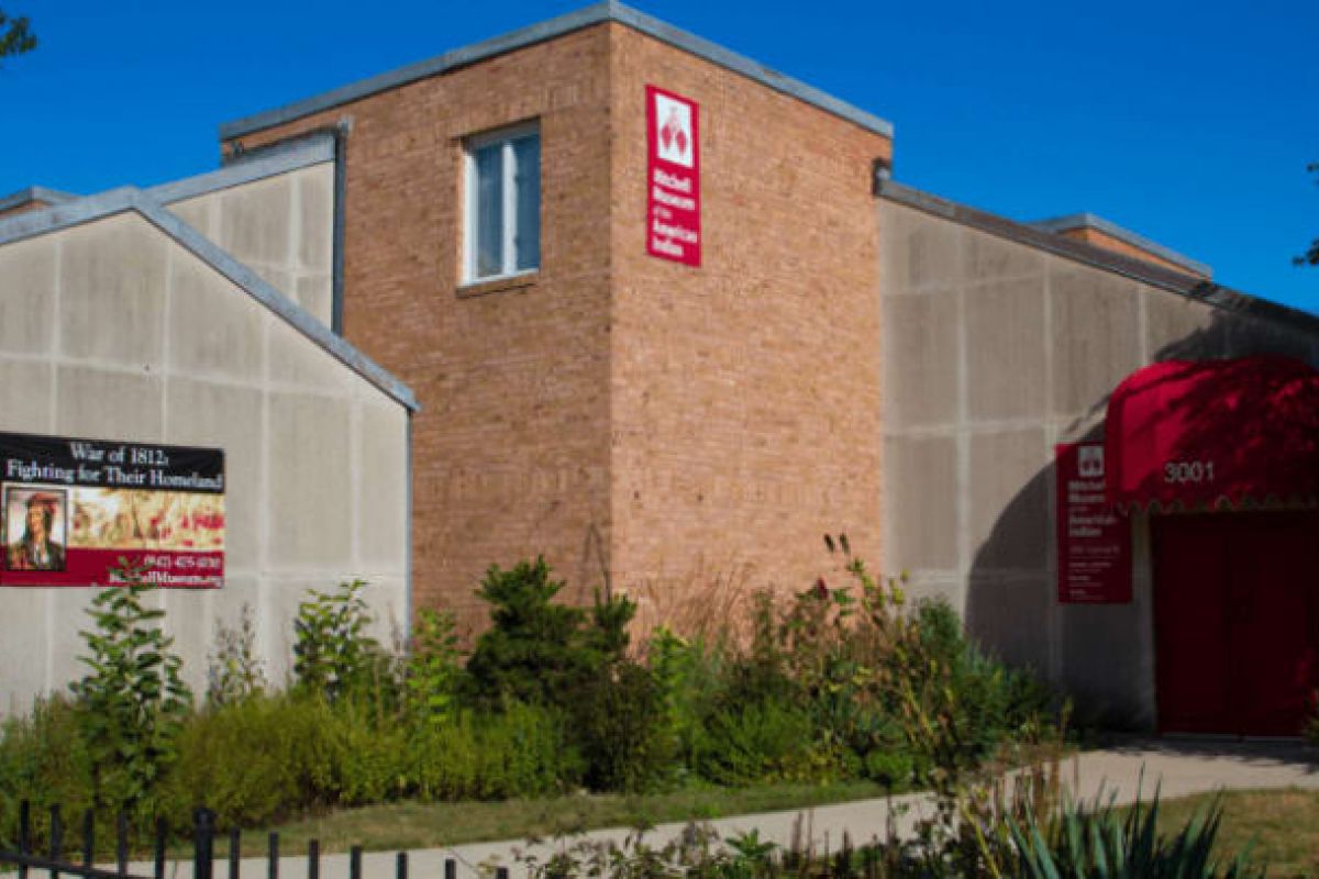 Optima Verdana luxury apartment with A tan and brick Mitchell Museum building with a red awning, sign, and greenery in front on a sunny day.