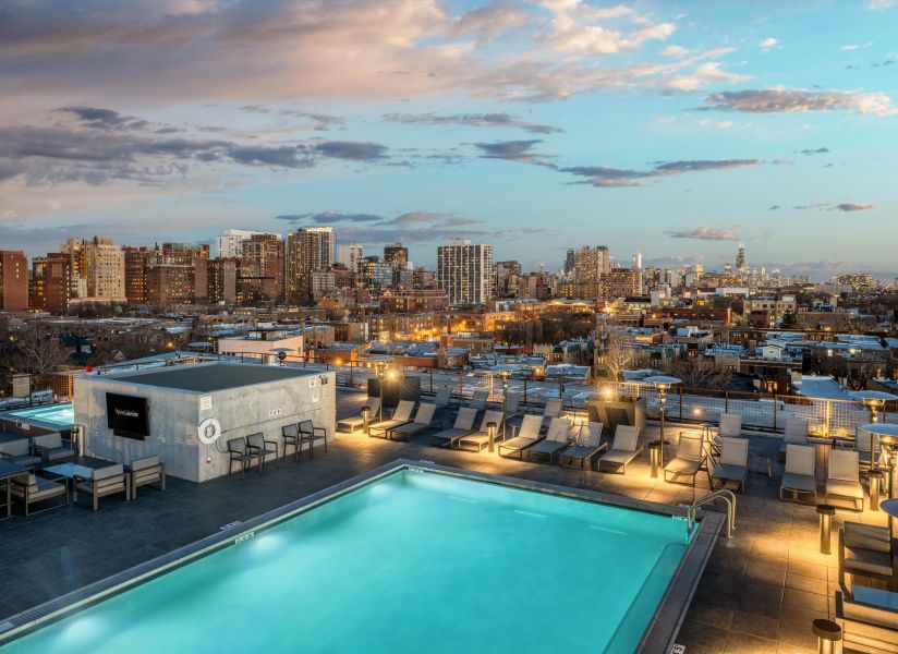 Rooftop pool and lounge area at Optima Lakeview apartments overlooking a city skyline at sunset, with scattered clouds in the sky.
