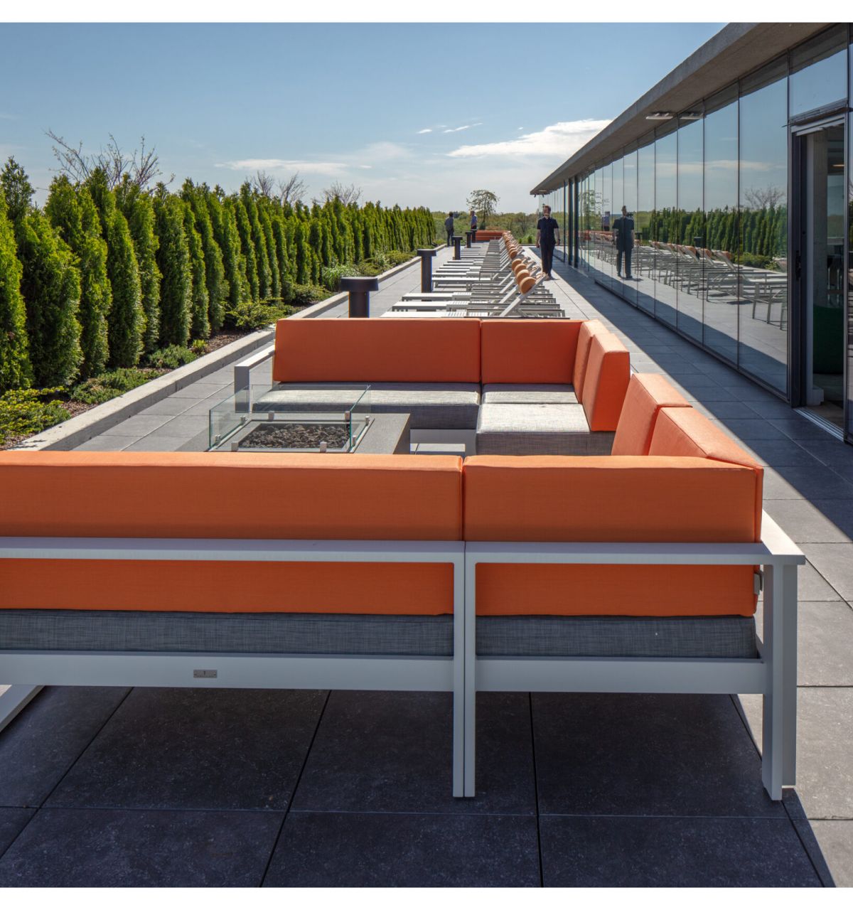 Optima Verdana luxury apartment with Outdoor rooftop patio with an orange sectional sofa, glass fire pit, lounge chairs, and greenery along the edge, under a clear sky.