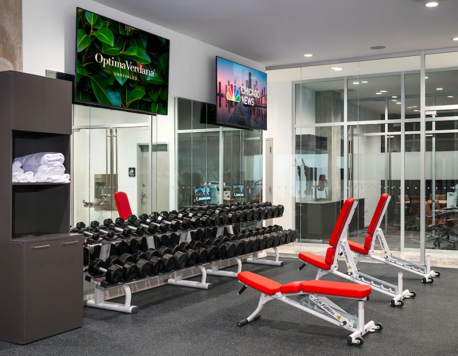 Optima Verdana luxury apartment with Modern gym with dumbbells, red benches, towels on shelves, and TVs on the wall in front of glass windows.