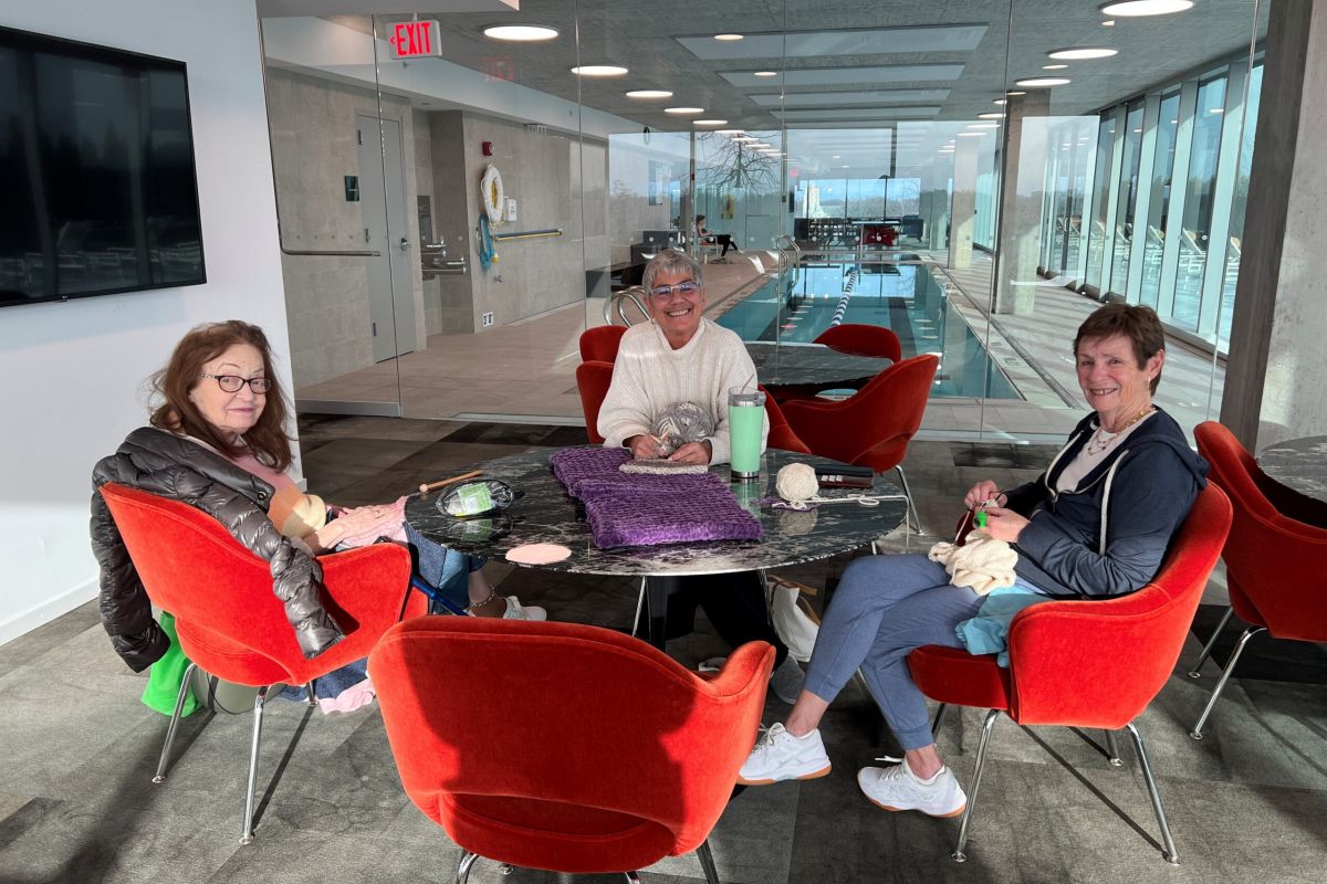 Optima Verdana luxury apartment with Three women sit and knit around a table in a bright lounge area at Optima Communities, with red chairs and a pool in the background.