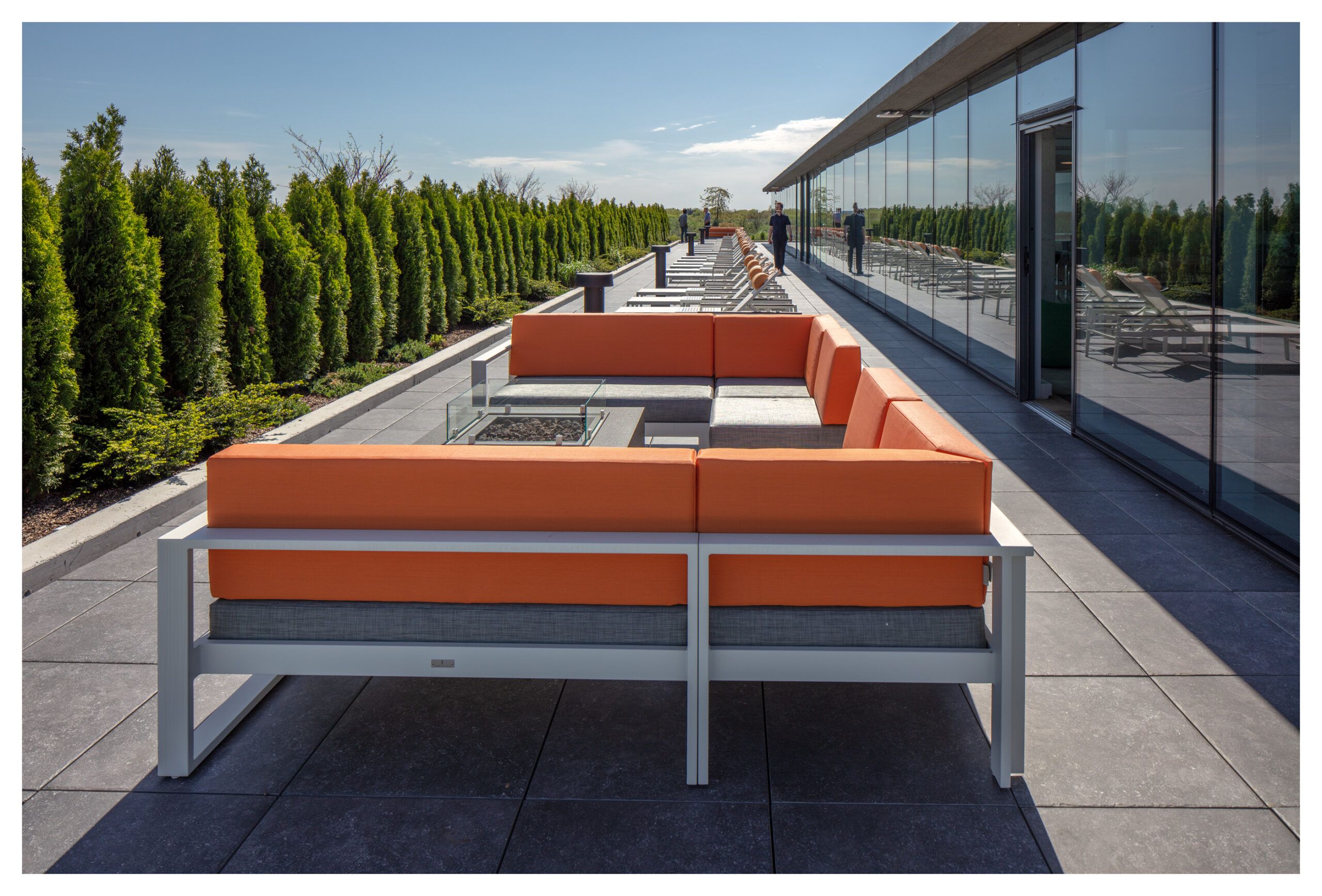 Optima Verdana luxury apartment with Outdoor rooftop patio with an orange sectional sofa, glass fire pit, lounge chairs, and greenery along the edge, under a clear sky.