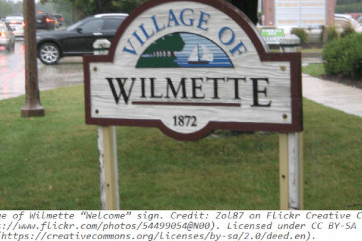 Optima Verdana luxury apartment with A wooden "Village of Wilmette" welcome sign with a sailboat and trees stands on the lakefront, revealing hidden geographies on a rainy day.