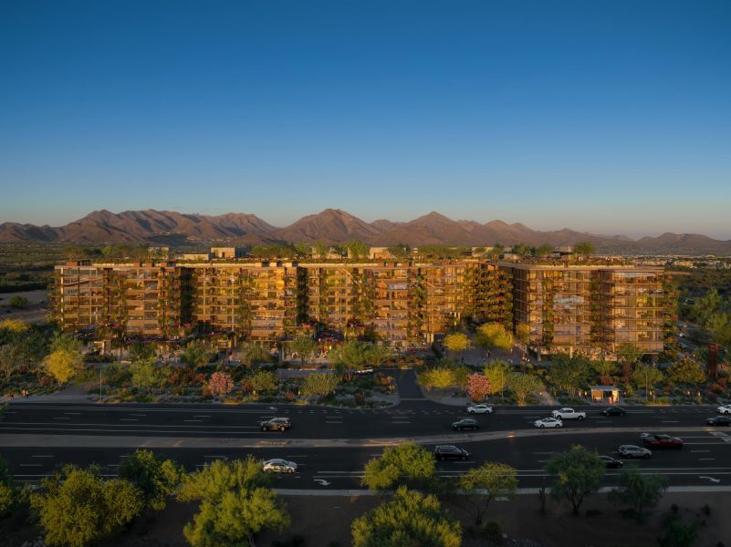Aerial view of modern multi-story buildings at Optima McDowell Mountain apartments surrounded by trees, with mountains in the background and a road in the foreground.