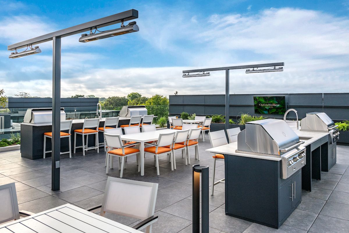 Optima Verdana luxury apartment with Rooftop patio with grills, dining tables, orange-cushioned chairs, and overhead heaters under a blue sky.
