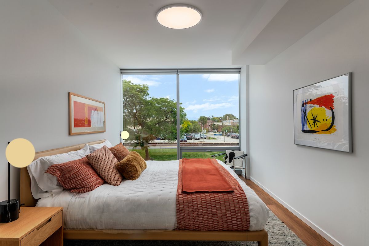 Optima Verdana luxury apartment with Modern bedroom with large window, white walls, wooden floor, and orange accents on bed and artwork.