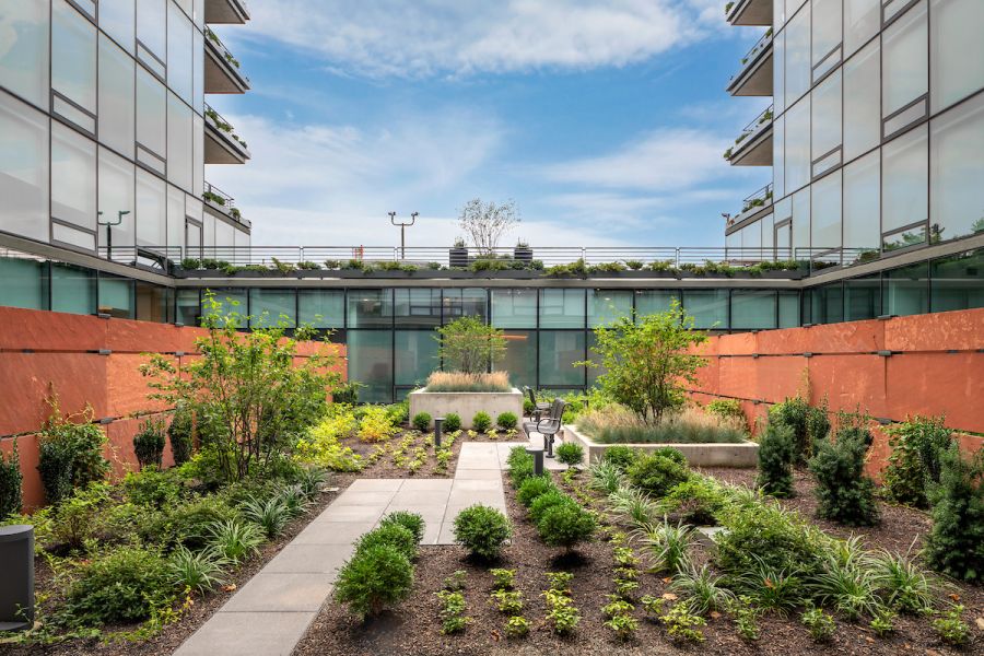 Optima Verdana luxury apartment with Modern courtyard garden with green plants, pathways, and glass buildings on both sides under a blue sky.