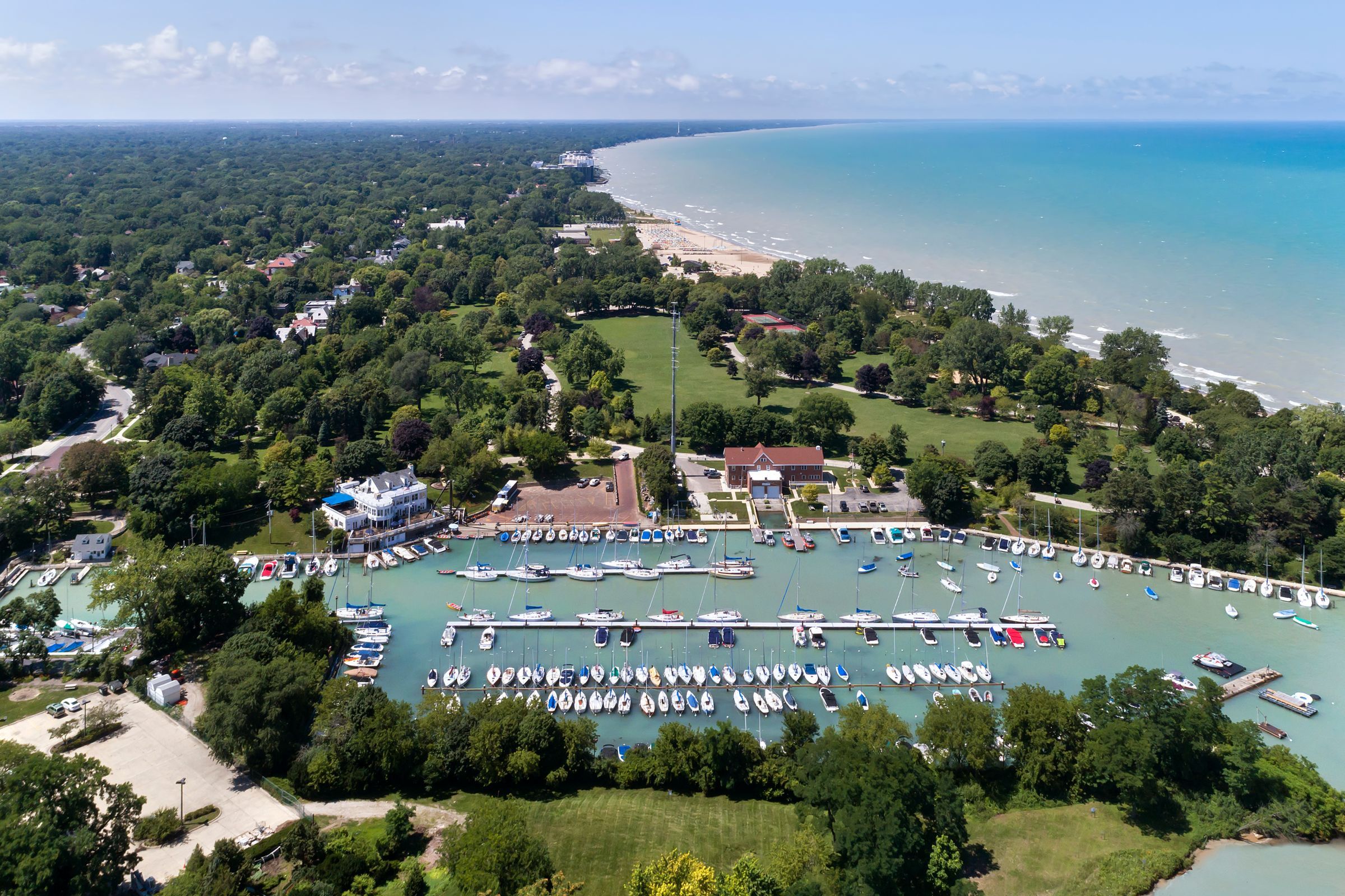Optima Verdana luxury apartment with Aerial view of a marina with many boats, a shoreline, and green trees along Lake Michigan on a sunny day.