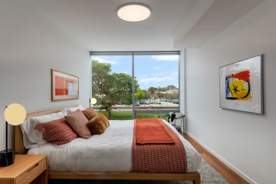 Optima Verdana luxury apartment with Modern bedroom with large window, white walls, wooden floor, and orange accents on bed and artwork.