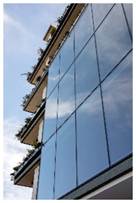 Optima Verdana luxury apartment with Tall glass building with balconies and plants, reflecting the blue sky and clouds.