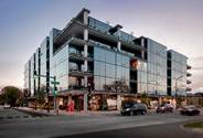 Optima Verdana luxury apartment with Modern glass building with balconies on a city corner, cars and pedestrians visible at street level.