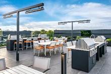 Optima Verdana luxury apartment with Rooftop patio with dining tables, chairs, and stainless steel grills under a blue sky with clouds.