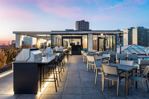 Optima Verdana luxury apartment with Rooftop patio with tables, chairs, and grills at sunset, overlooking city buildings under a colorful sky.
