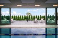 Optima Verdana luxury apartment with Indoor pool with open glass doors leading to an outdoor patio with lounge chairs and trees under a blue sky.